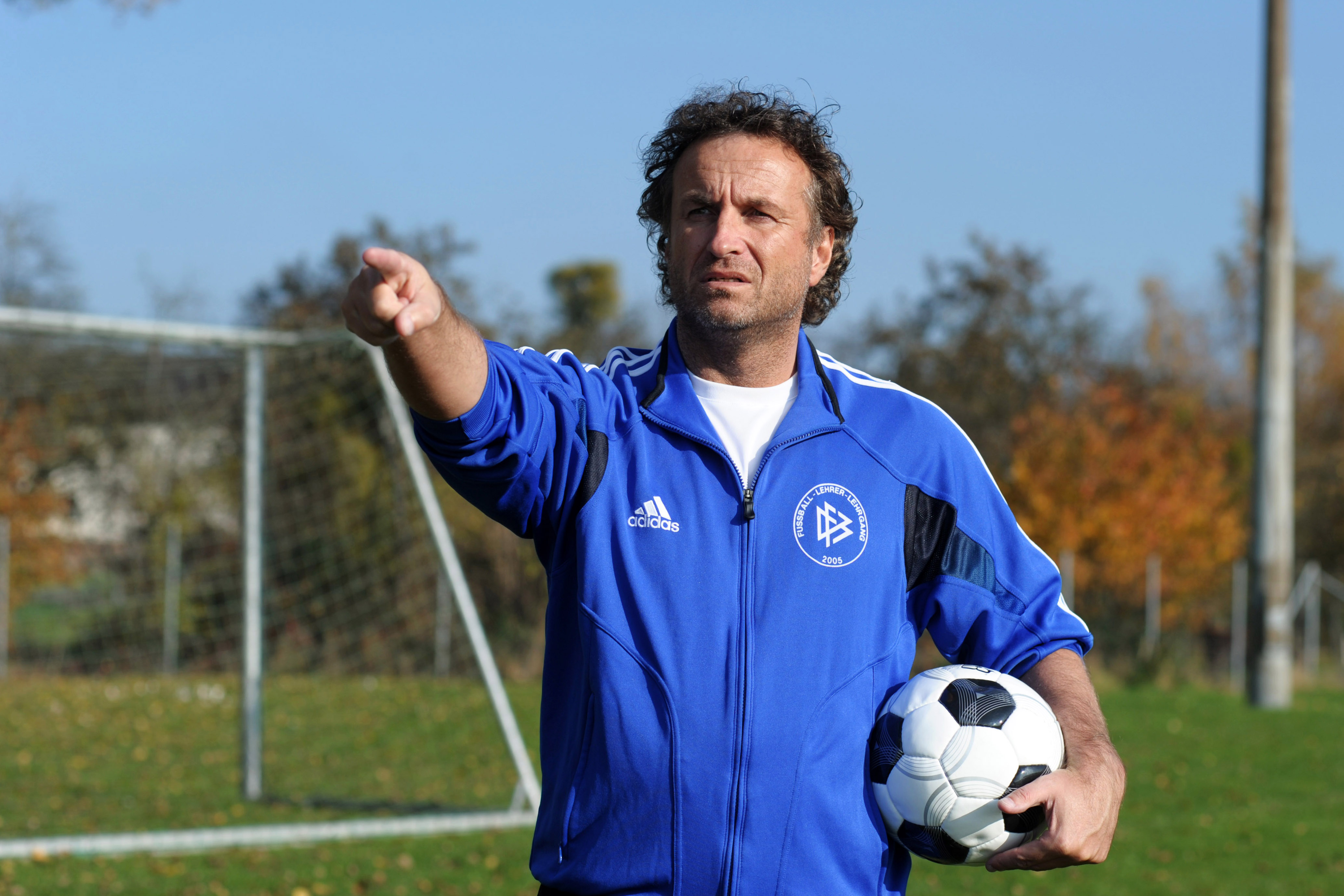 Alfred Kaminski gibt auf dem Trainingsplatz eine Anweisung, mit einem Fußball unter dem Arm.